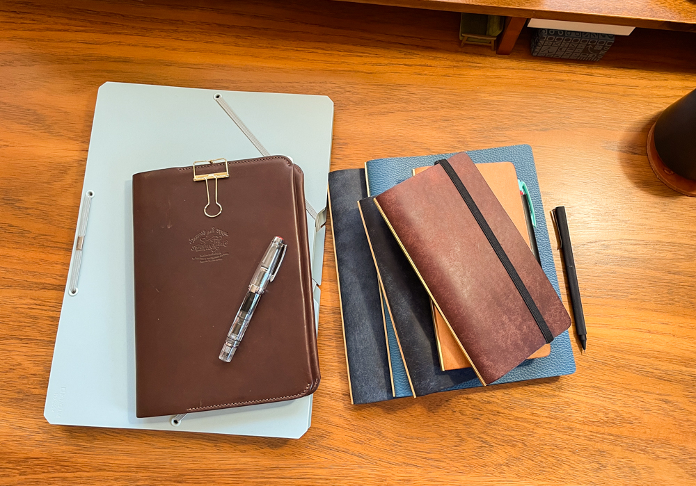 Desk view of a stack of PLOTTER leather binders and a brown leather notebook.
