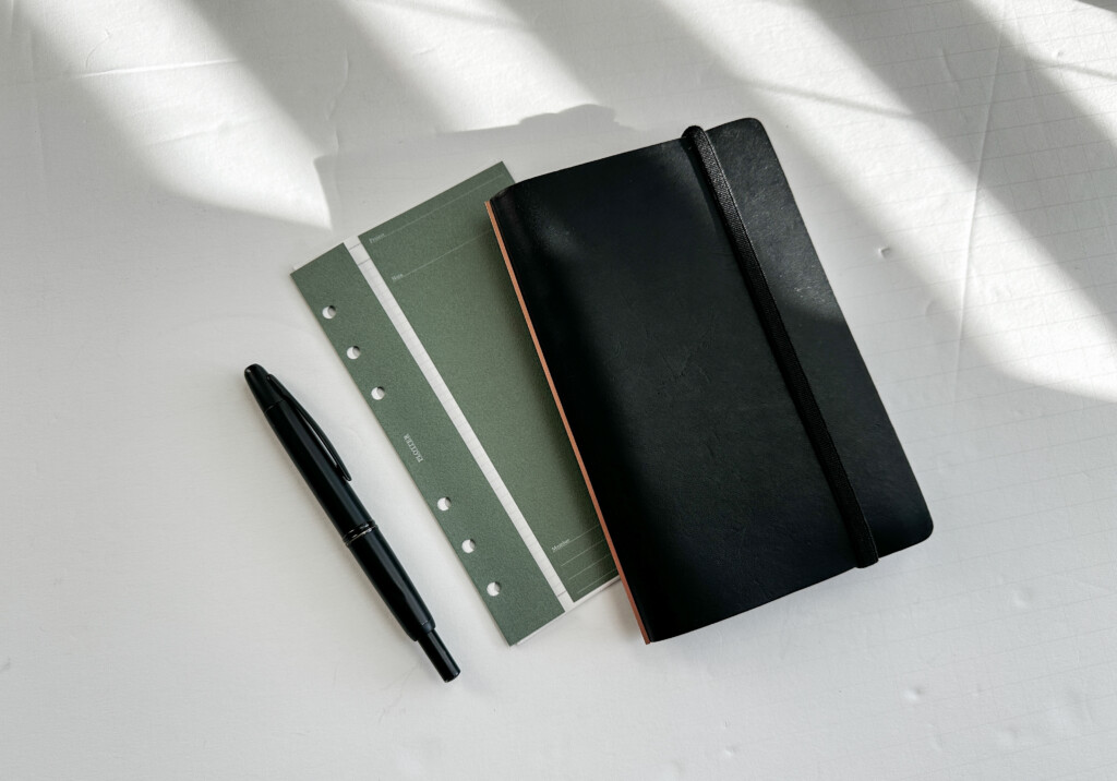 PLOTTER Bible Size Leather Binder in Black with a pen and a green paper folder.