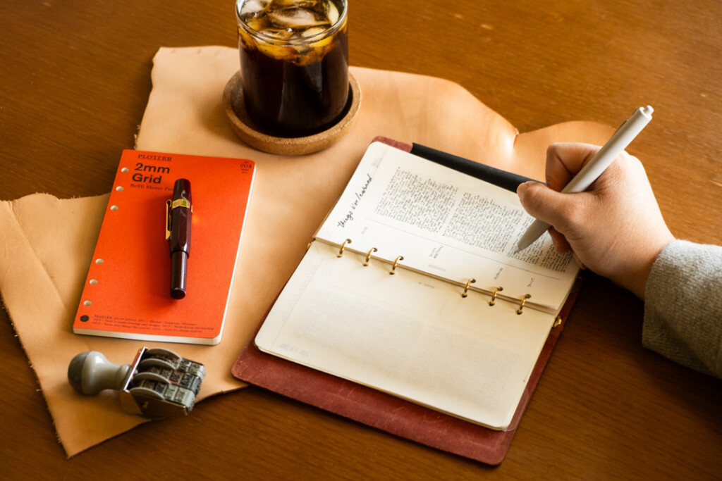 Person writing in a PLOTTER Leather Binder open to a page of notes called "things I learned this year" beside ice coffee, Refill Memo Pad, Pen and eye glasses.