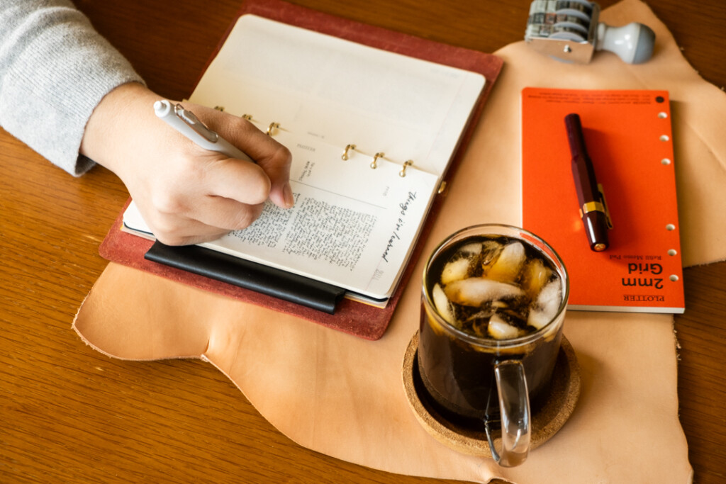 Person writing in a PLOTTER Leather Binder open to a page of notes called "things I learned this year" beside ice coffee, Refill Memo Pad, Pen and eye glasses.