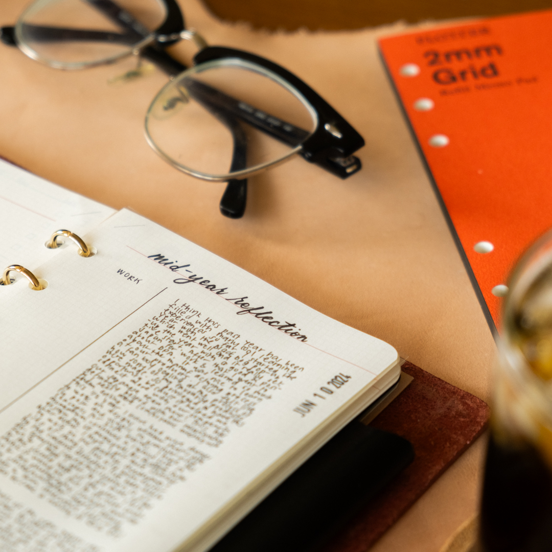 Close-up of PLOTTER Leather Binder open with notes beside ice coffee, Refill Memo Pad, Pen and eye glasses.