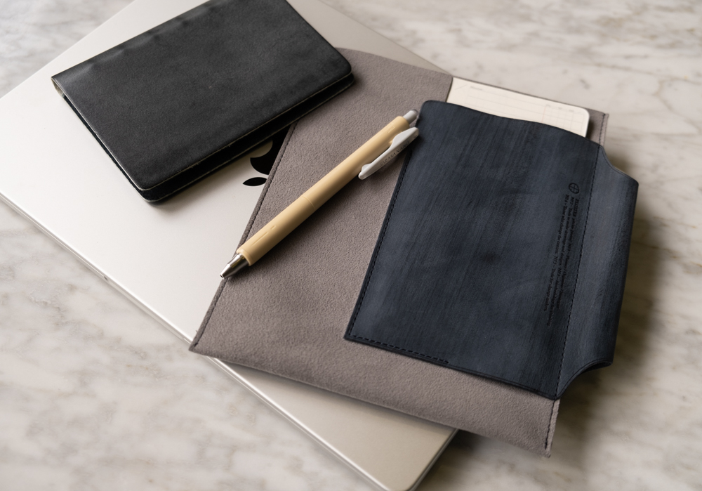 A Gray x Navy PLOTTER Leather Binder Case positioned with a pen and a PLOTTER in Mini Size.
