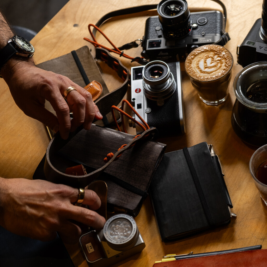 Man organizing cameras, PLOTTER leather binders and coffee cups on wooden table.