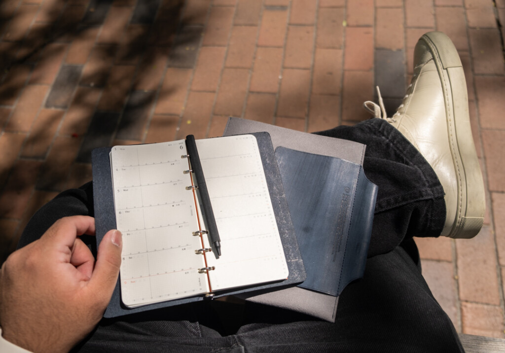 Narrow Size PLOTTER Leather Binder and Leather Binder Case in Gray and Blue on a man's lap who is sitting on a park bench.