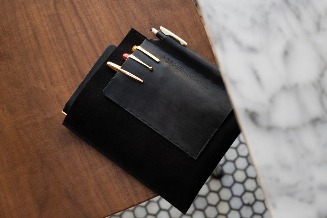 PLOTTER Leather Binder Case in Bible Size in Black which has fountain pens stored on the front leather pocket, on top of a wooden chair.