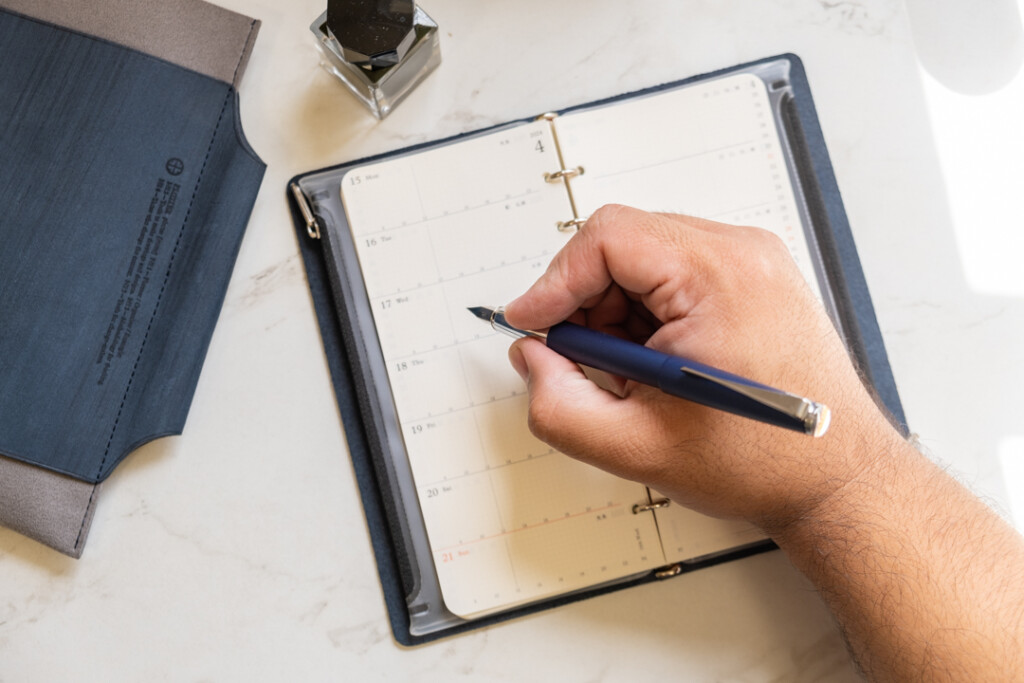 Over-head view of a person writing in an PLOTTER Narrow Size Leather Binder with a blue fountain pen.