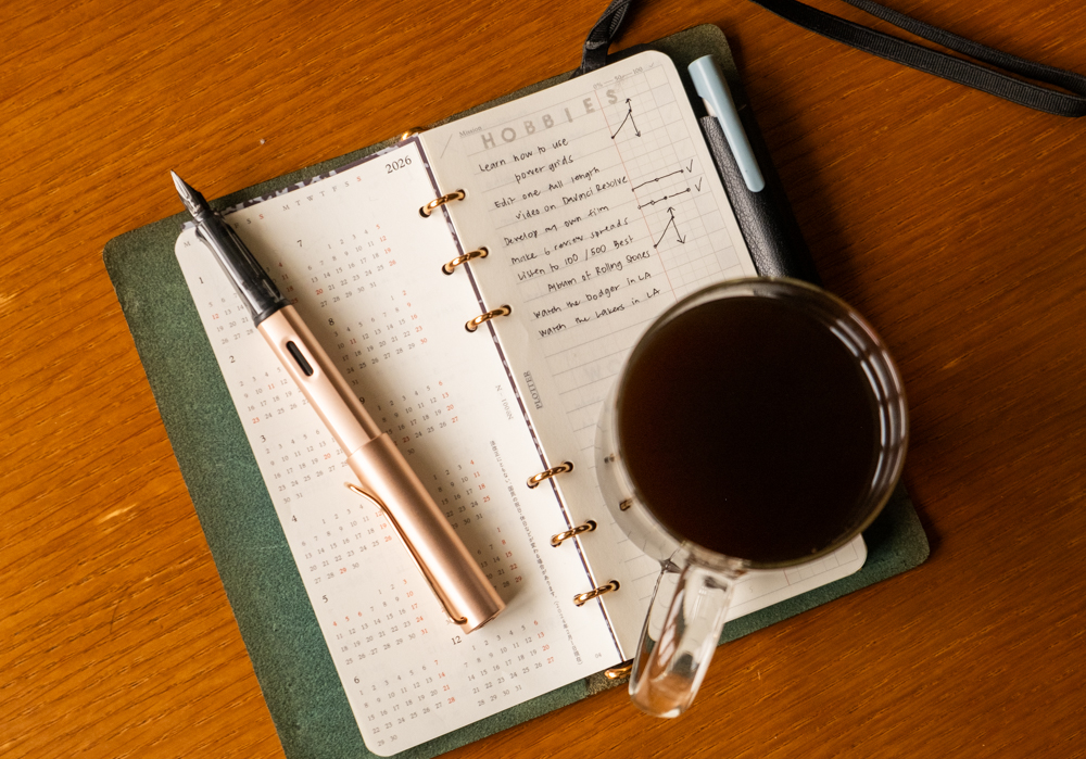 A PLOTTER Narrow Size Leather Binder open to a list of new year's goals related to hobbies. There is a fountain pen and a coffee near the PLOTTER.