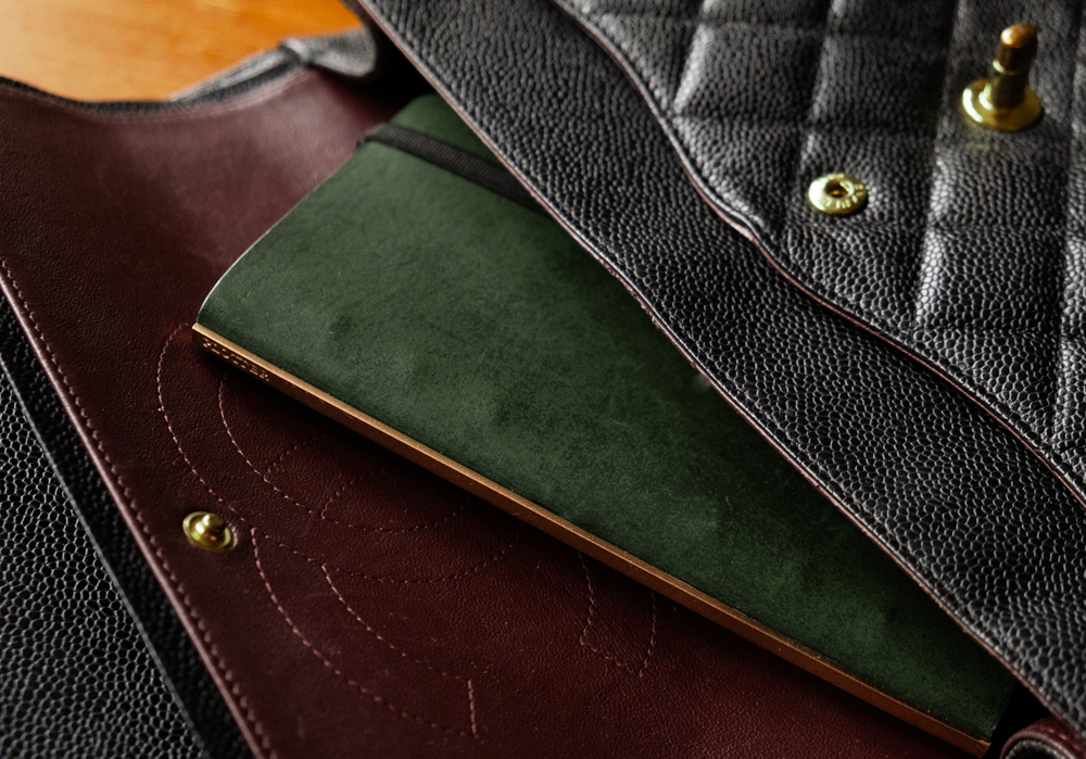 A close up of a green PLOTTER Narrow Size Leather Binder inside a black leather hand bag.