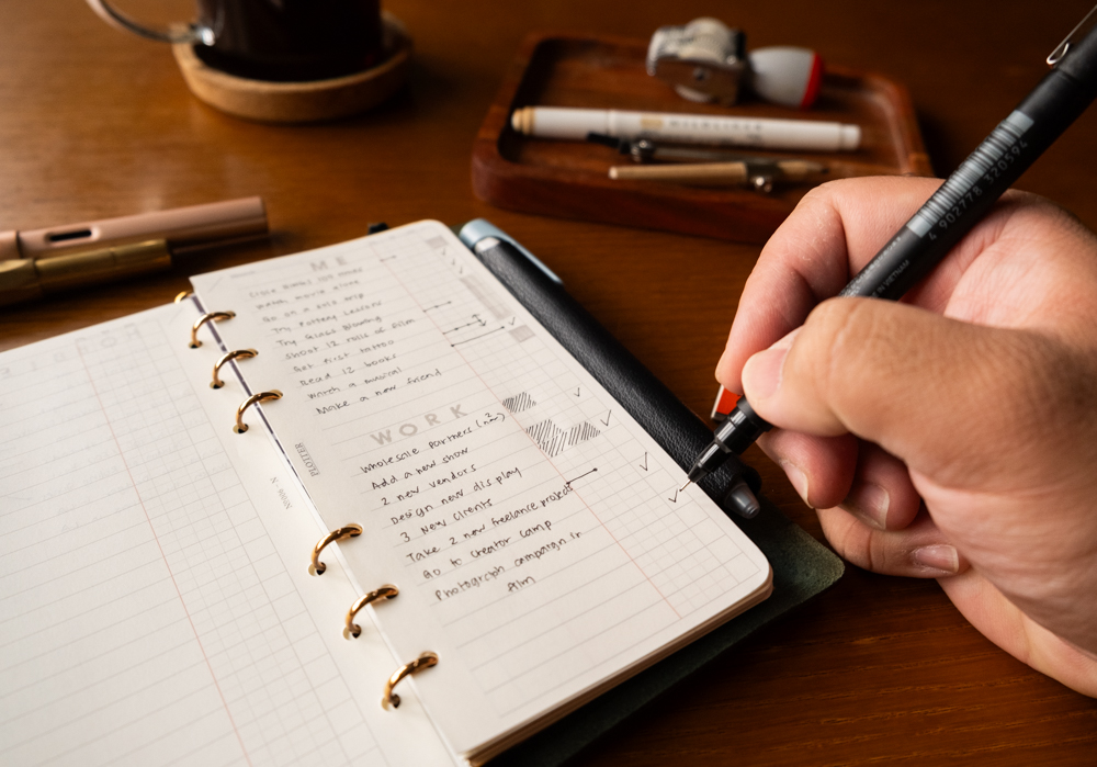 A person writing on PLOTTER Narrow Size Leather Binder open to a List of new year's work and personal goals.