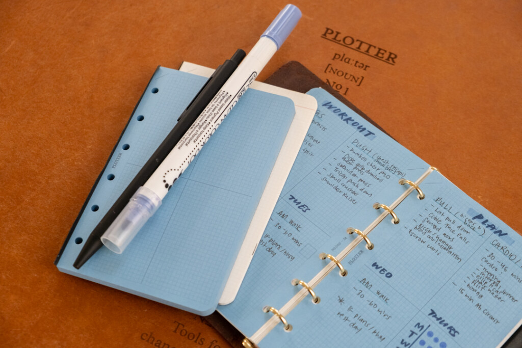 PLOTTER Mini Size open to a spread with a workout routine with a pens, markers and PLOTTER Refill Memo Pads beside it.